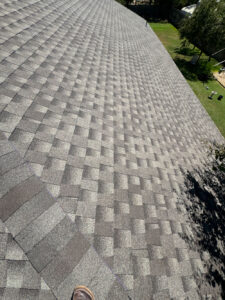 A view of a large, sloped roof covered in gray shingles in a checkered pattern. A small section of a shoe is visible at the bottom edge of the image. The surrounding area includes trees and grass.