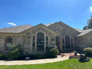 A single-story house with a stone exterior and arched windows under a clear blue sky. A well-maintained lawn surrounds the walkway leading to the entrance. Gardening tools and equipment are visible near the front porch.