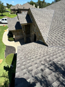 Aerial view of a stone house with a steep, shingled roof on a sunny day. A white van is parked on the driveway, surrounded by green lawns and trimmed shrubs. Shadows are visible on the roof and grass.