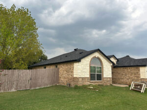 Brick house with a mix of tan and white exterior, featuring a large arched window and a dark shingled roof. In the background, cloudy skies loom over the house. A tall wooden fence and a patch of green grass are visible in the foreground.