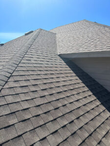 Close-up view of a house roof with gray asphalt shingles under a clear blue sky. The roof features intersecting lines and angles, showcasing the structure and pattern of the shingles. Bright sunlight casts shadows across the surface.