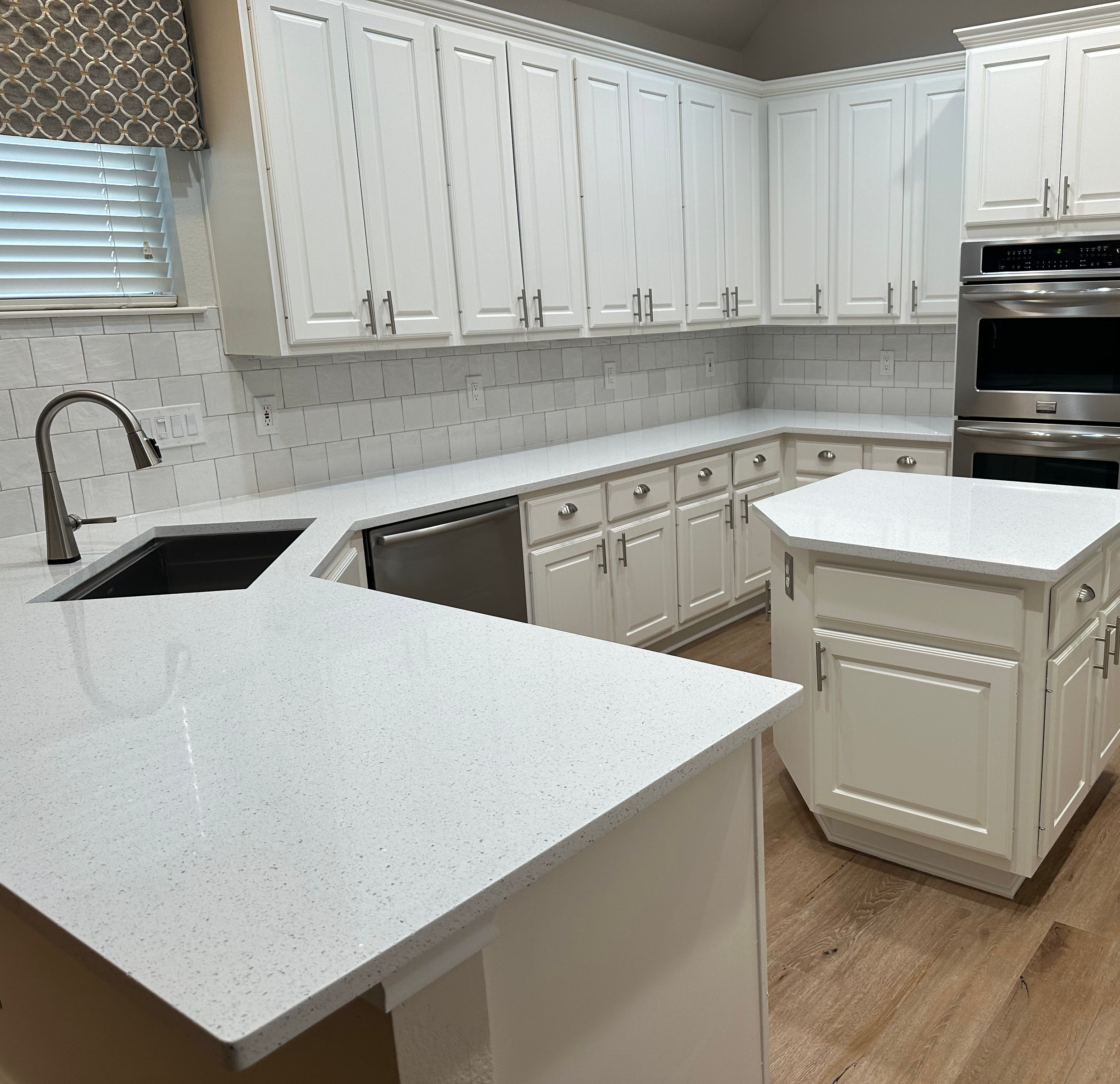 Modern kitchen with white cabinetry and countertops, featuring an island and stainless steel appliances. A window with a patterned valance is above the sink, and the floor is wooden. The overall design is sleek and bright.
